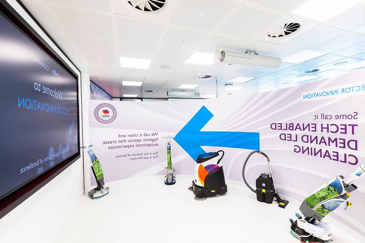 A collection of five cleaning robots and equipment at the Mitie 清洁 & 卓越卫生中心. The wall behind has text of 'Some call it 科技 enabled demand led cleaning'